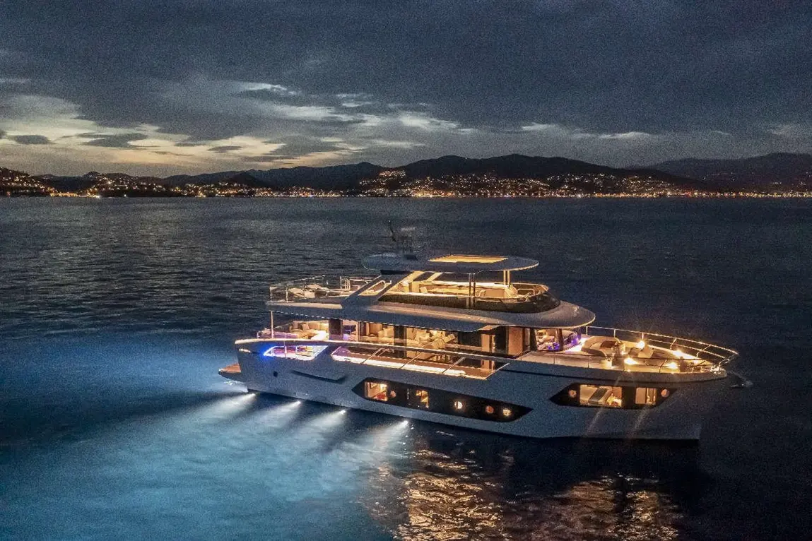 Luxury yacht illuminated at night, gliding on calm waters with distant city lights and mountains in the background.